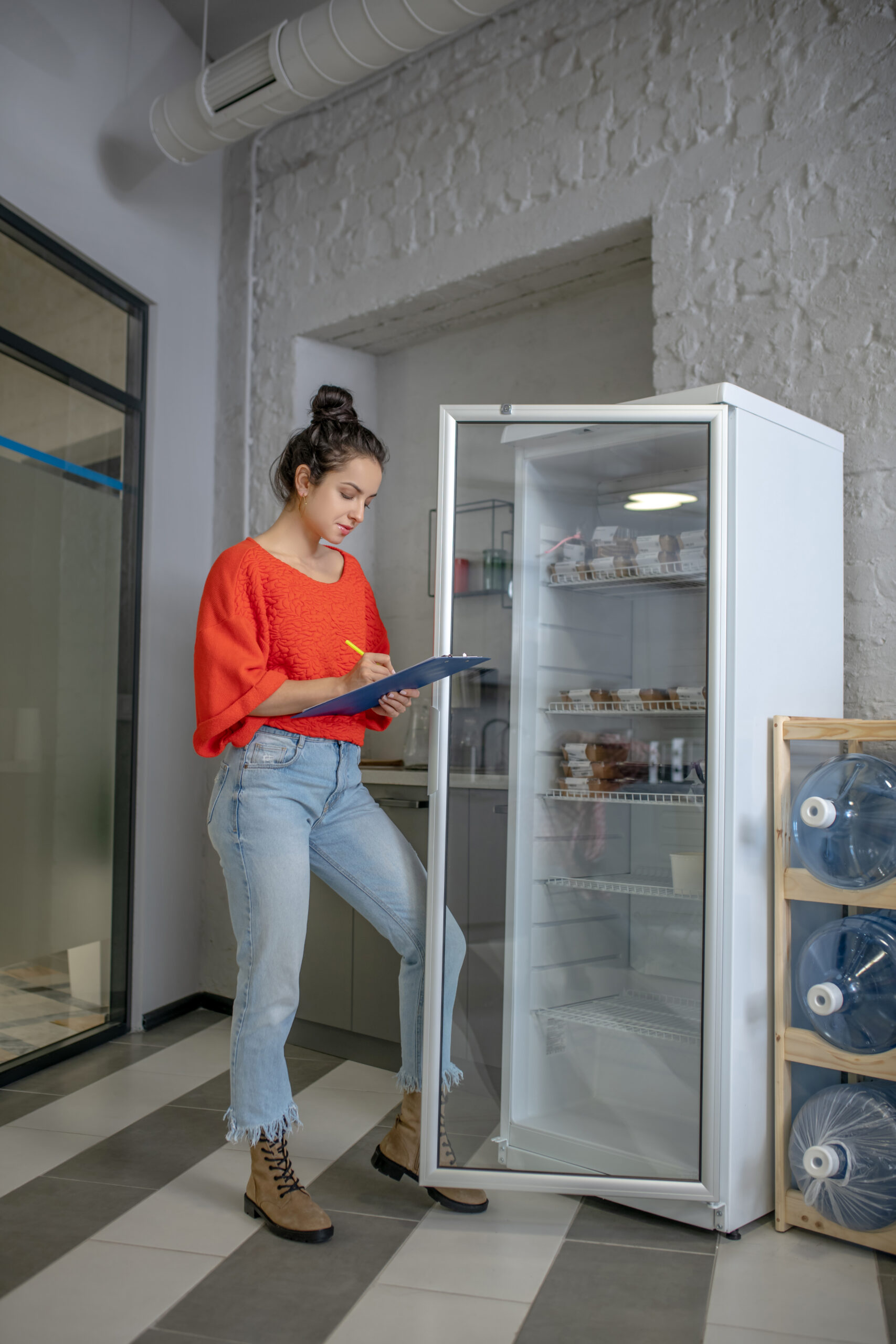 Indoors. Young woman holding fridge door with leg, making notes, counting lunch boxes